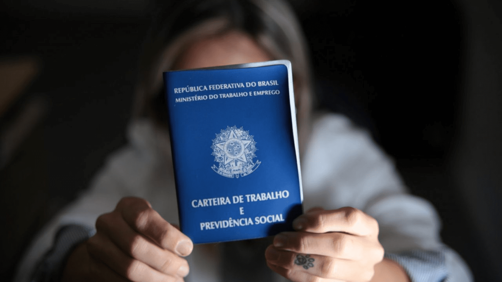 Imagem de pessoa desfocada segurando na frente do rosto uma Carteira de Trabalho