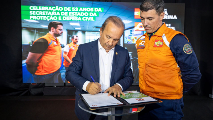 Governador de SC Jorginho Melo assinando decreto ao lado de agente da Defesa Civil