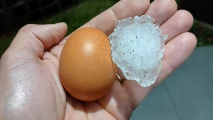 Mão segurando um ovo de galinha e pedra de gelo (granizo) do tamanho do ovo
