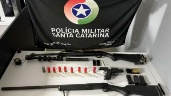 Bandeira da Polícia Militar ao fundoe armas e munições sobre uma mesa apreendidas em Abelardo Luz
