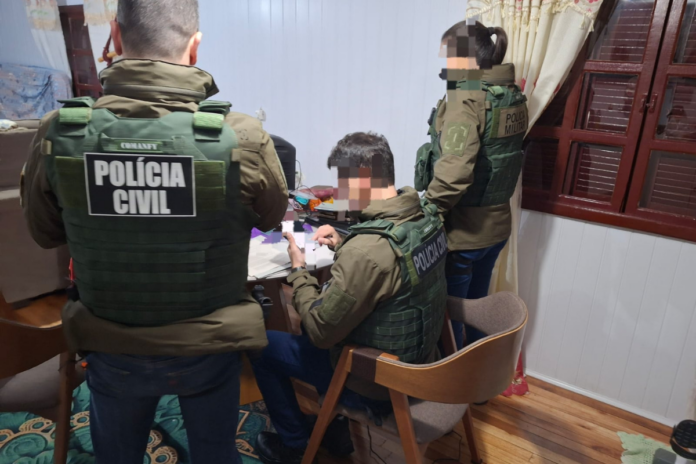 Três policiais civis fardados em uma sala, com um deles sentado em uma cadeira