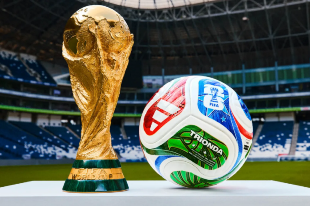 Troféu da Copa do Mundo da FIFA ao lado de uma bola de futebol sobre uma mesa branca em frente á um estádio