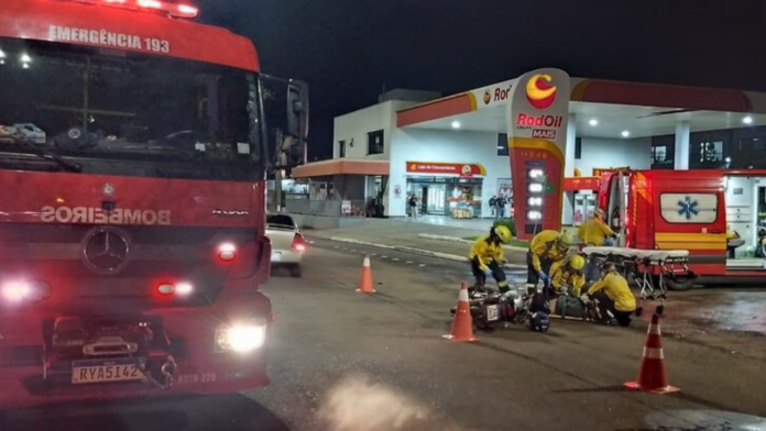 Carro dos bombeiros parado na rua e socorristas dando atendimento á motoqueiro caido após bater em caminhonete.